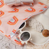 A pink reusable waterproof baby wet bag with a rainbow pattern and a pair of light-colored sunglasses resting on a fluffy whi