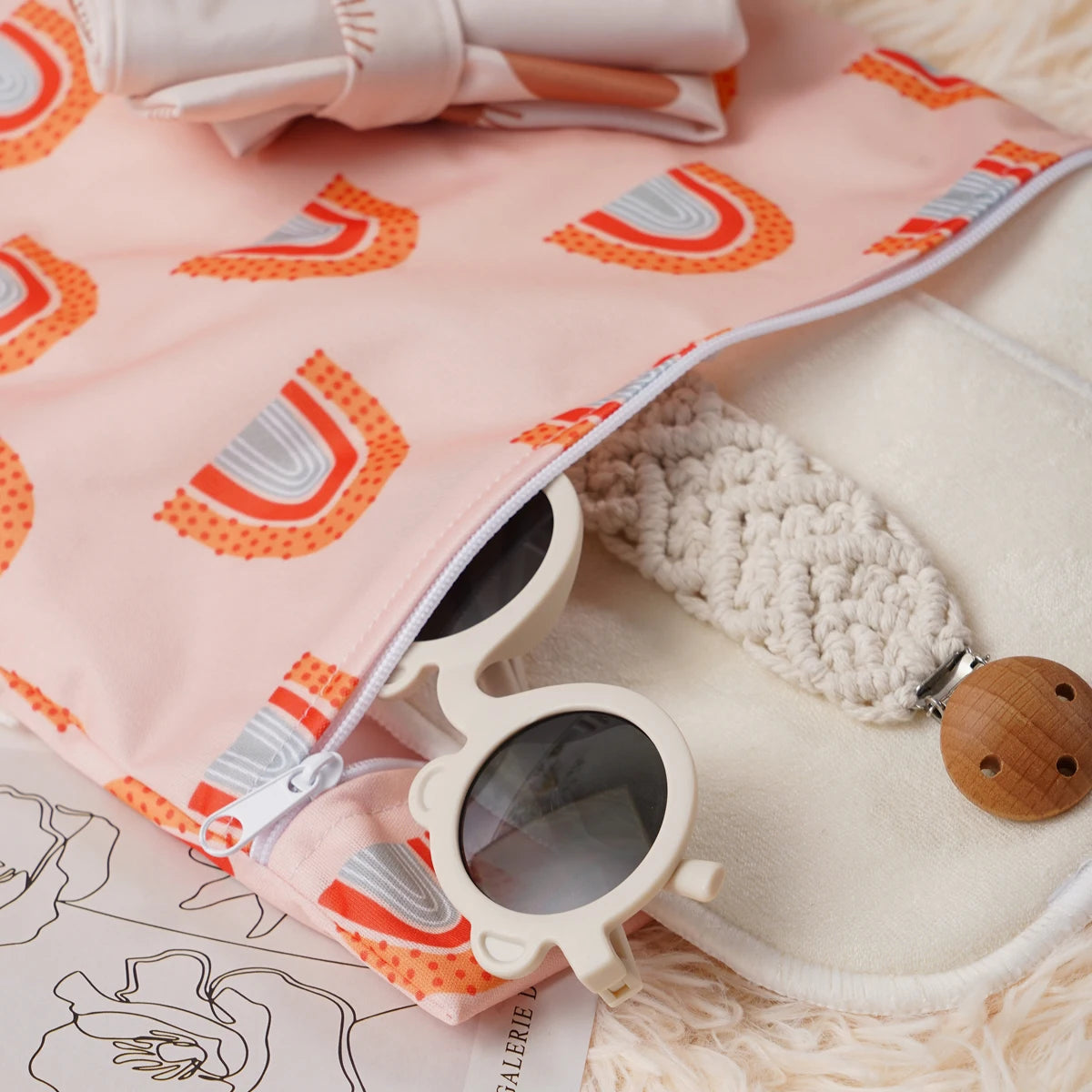 A pink reusable waterproof baby wet bag with a rainbow pattern and a pair of light-colored sunglasses resting on a fluffy whi
