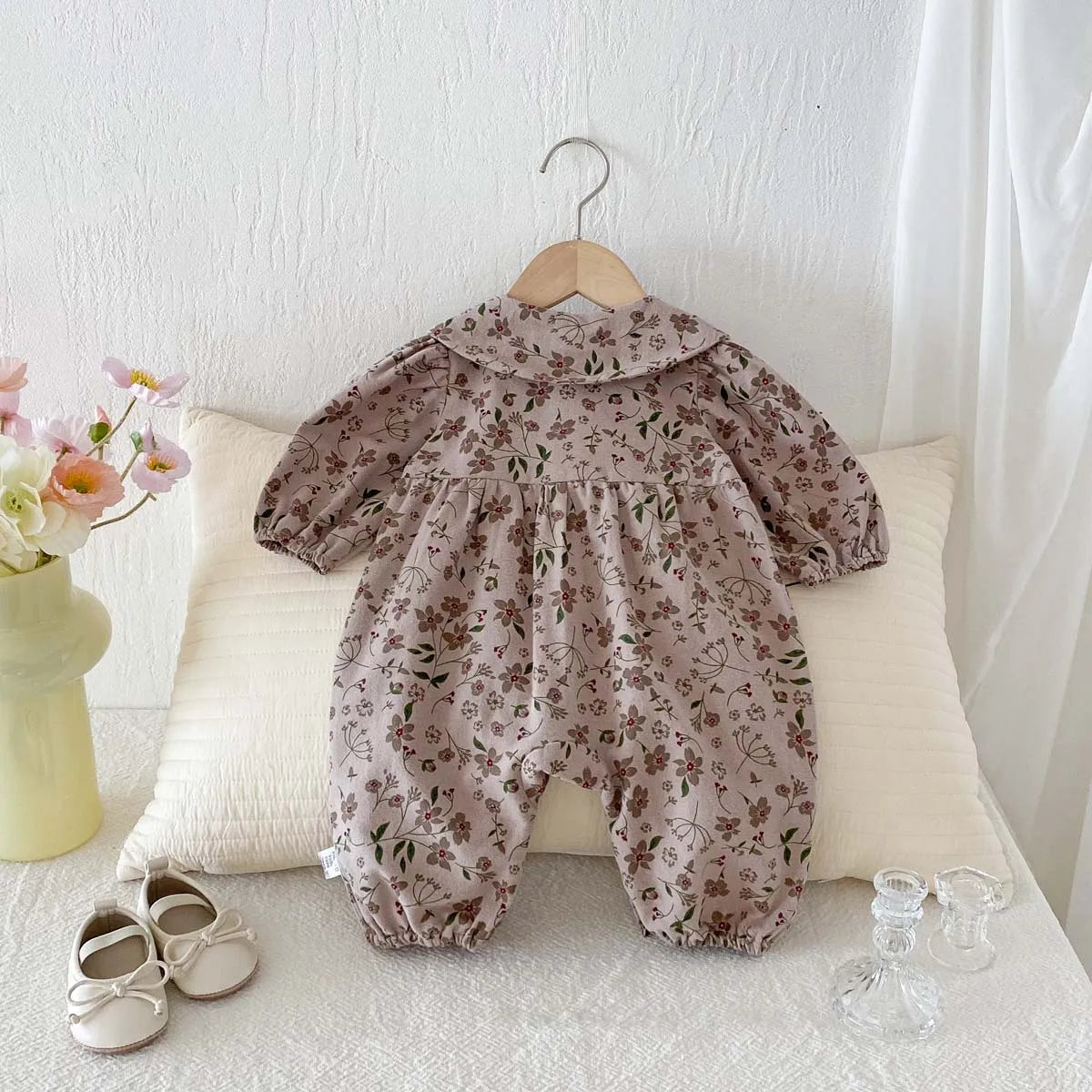 A floral doll collar baby girl long sleeve jumpsuit is displayed on a white surface with a light colored pillow.