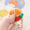 A hand holds an orange crochet fruit keychain pendant with white stripes and a green charm against a light background.
