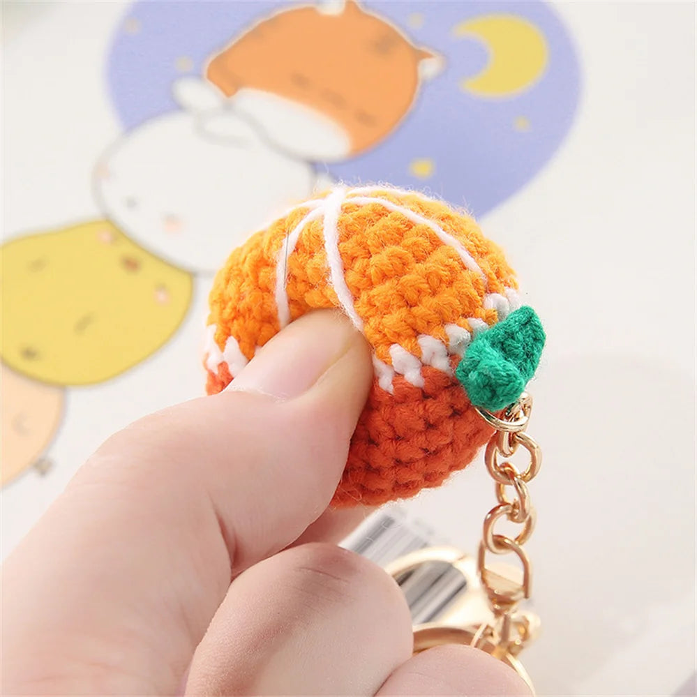 A hand holds an orange crochet fruit keychain pendant with white stripes and a green charm against a light background.