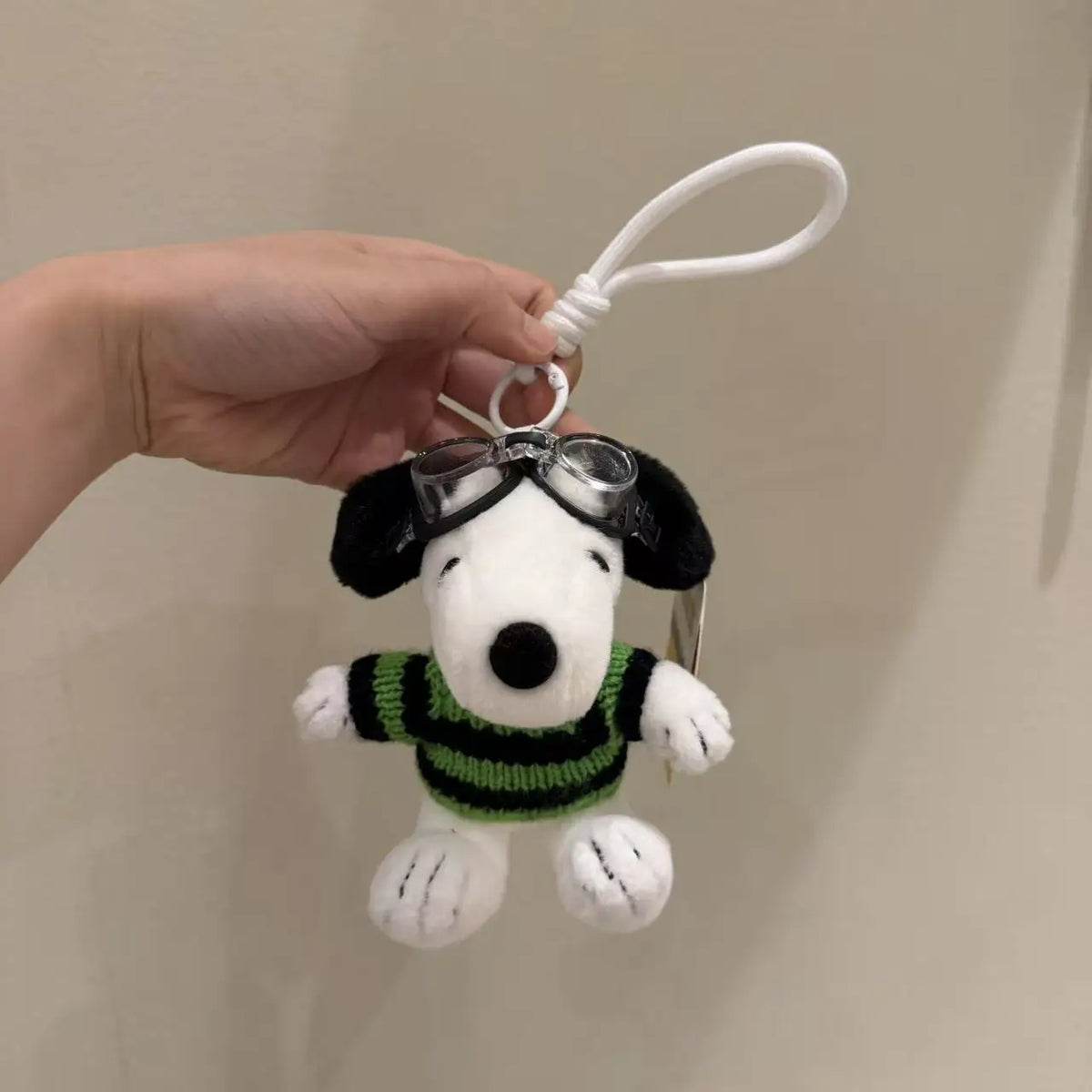 A white Snoopy plush toy with black ears and a green sweater is being held by a hand, attached to a white cord.