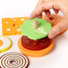 A hand is stacking wooden burger-themed toys, including a green top piece and a red base, onto an orange platform.