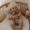 A baby wearing a beige knit hat, long-sleeve top, and matching pants sitting in a wooden crib.