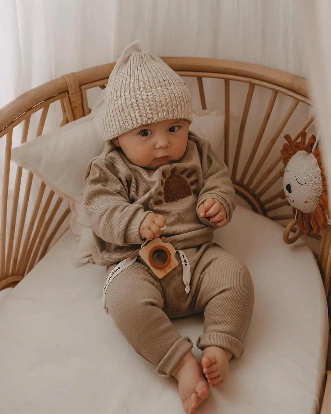 A baby wearing a beige knit hat, long-sleeve top, and matching pants sitting in a wooden crib.