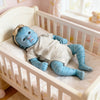 A handcrafted reborn baby doll with blue skin and light gray clothing lying in a white wooden crib.