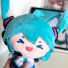 A close-up of a teal Hatsune Miku plush doll with black and pink accents being held against a blurred background.