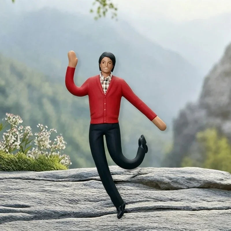 Une figurine en peluche à t-shirt rouge et pantalon noir se tient sur une surface rocheuse devant un arrière-plan montagneux 