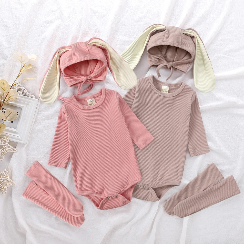 A pink and beige rabbit romper set for babies includes a matching hat and socks laid out on white fabric.