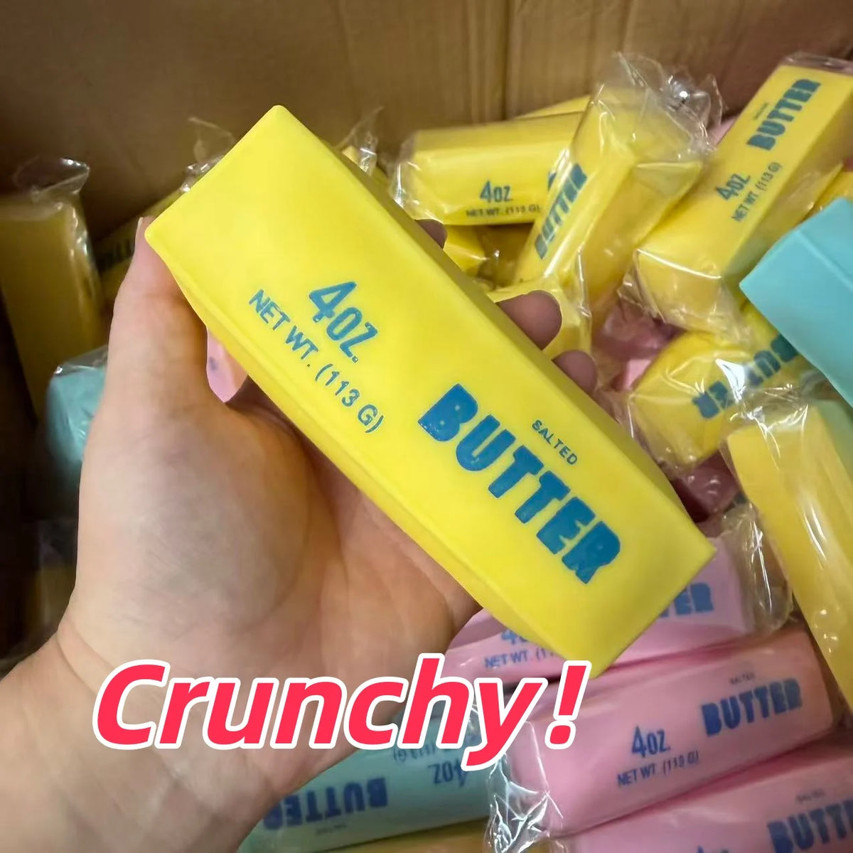 A hand holds a yellow butter stick squishy stress ball with salted butter visible in the background.