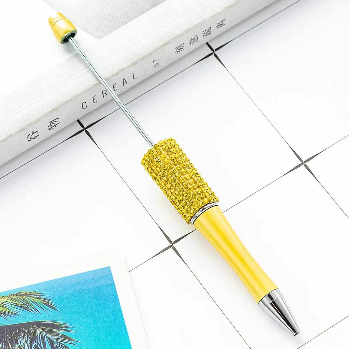 A gold and yellow beaded ballpoint pen craft kit with a needle is shown on a white surface.