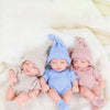 Three newborn simulation dolls wearing knitted outfits in soft pastel colors on a white fluffy surface.