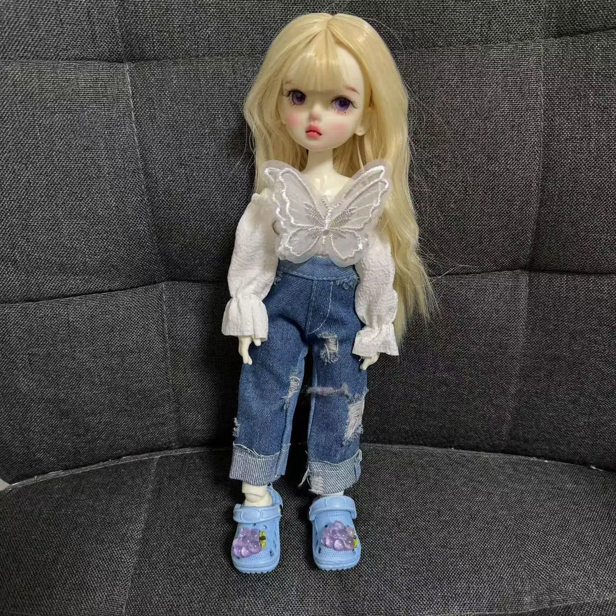 A blonde doll wearing a white shirt and denim jeans with butterfly wings, posing on a dark gray fabric background.