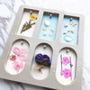 A silicone mold tray with six compartments containing various natural elements like flowers, stones, and botanicals for craft