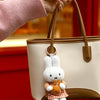 A beige tote bag with a brown strap and a white Miffy plush keychain charm is held by a hand against a red background.