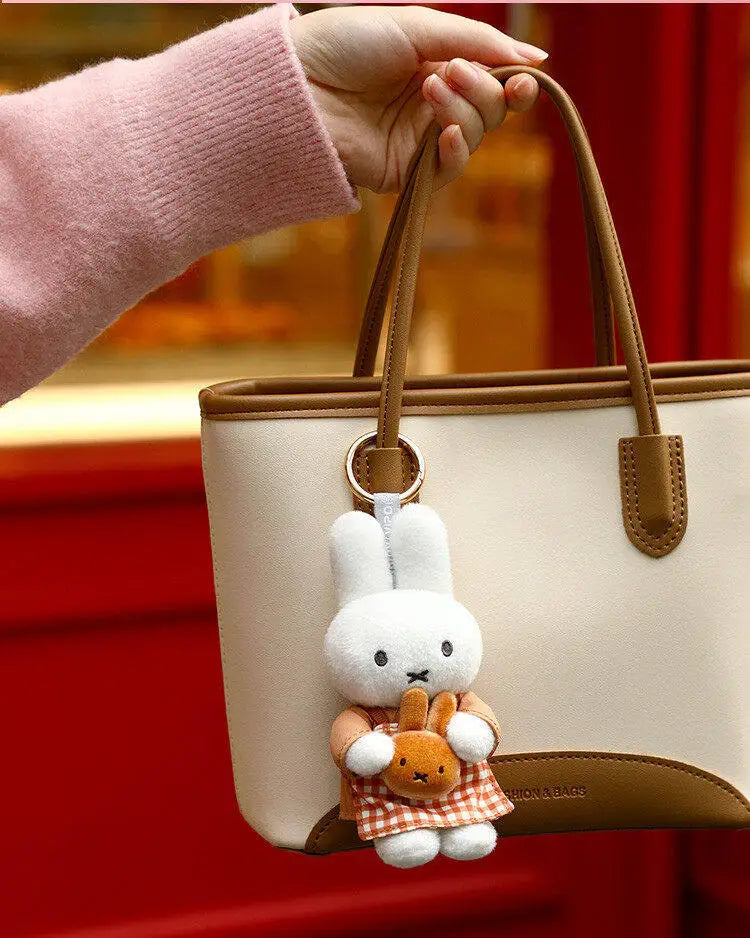 A beige tote bag with a brown strap and a white Miffy plush keychain charm is held by a hand against a red background.