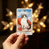 A hand holds a tarot card depicting The Empress, featuring a cat face and a red cloak against a blue background.