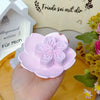 A hand holds a pink silicone flower mold for soap, candle, or craft making on a wooden surface.