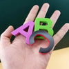 A hand holds three magnetic letters A, B, and a curved letter, all in purple, green, and black.