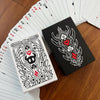A black and white poker card deck featuring intricate line art and red heart symbols, displayed next to a sealed box.