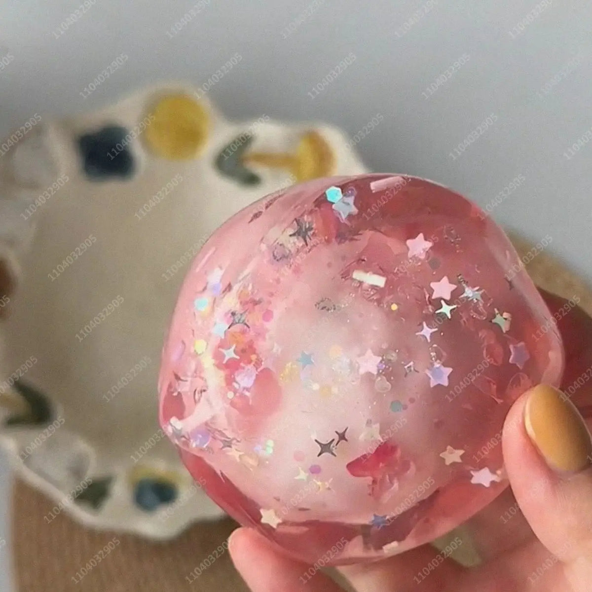 A hand holds a pink, translucent stress ball filled with glitter and small stars.