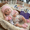 A realistic soft silicone reborn baby doll wearing a pink knitted hat and floral outfit, resting in a lace-lined basket.