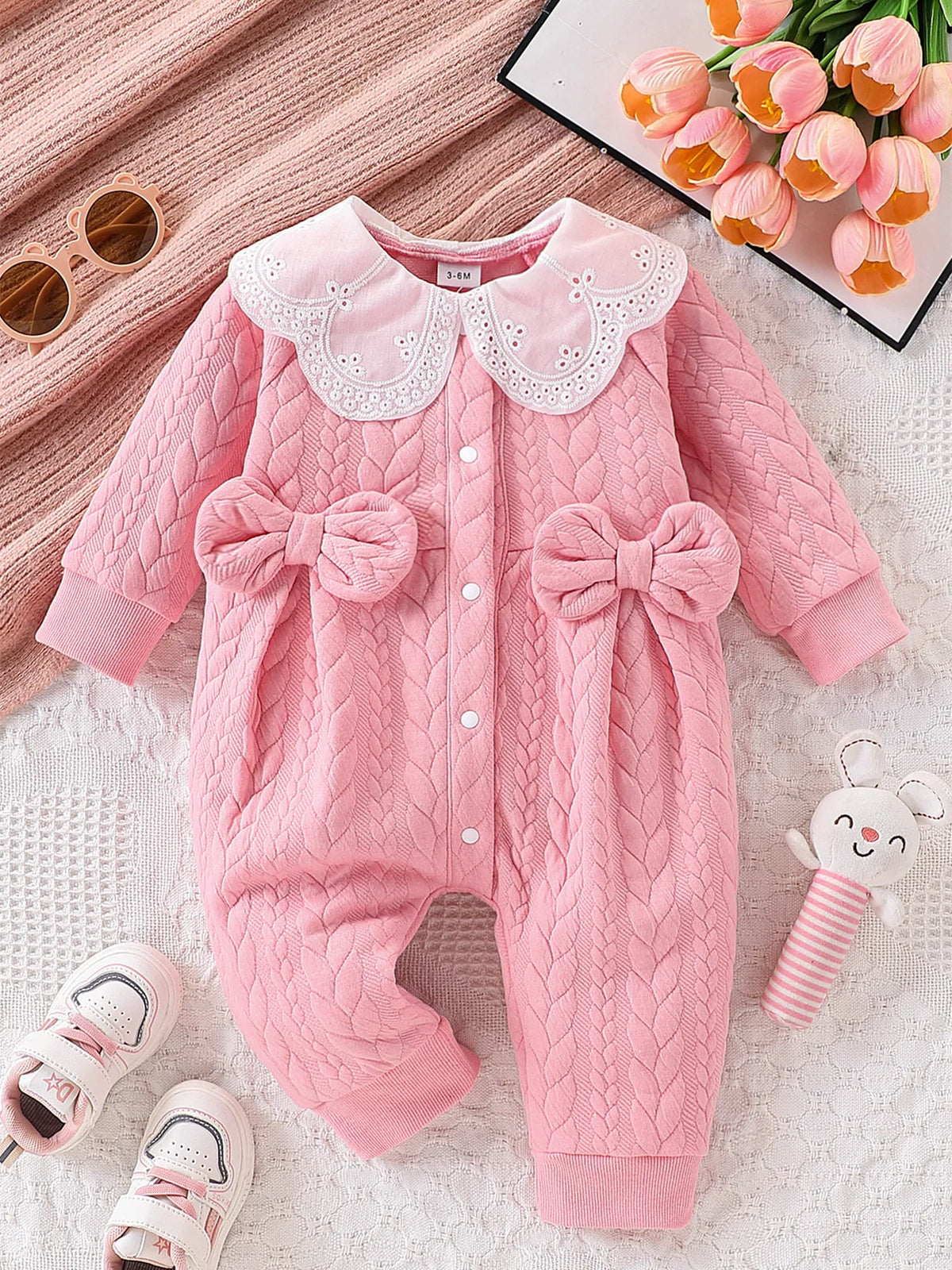 Pink quilted baby girl romper with a lace collar lying on a white textured surface.