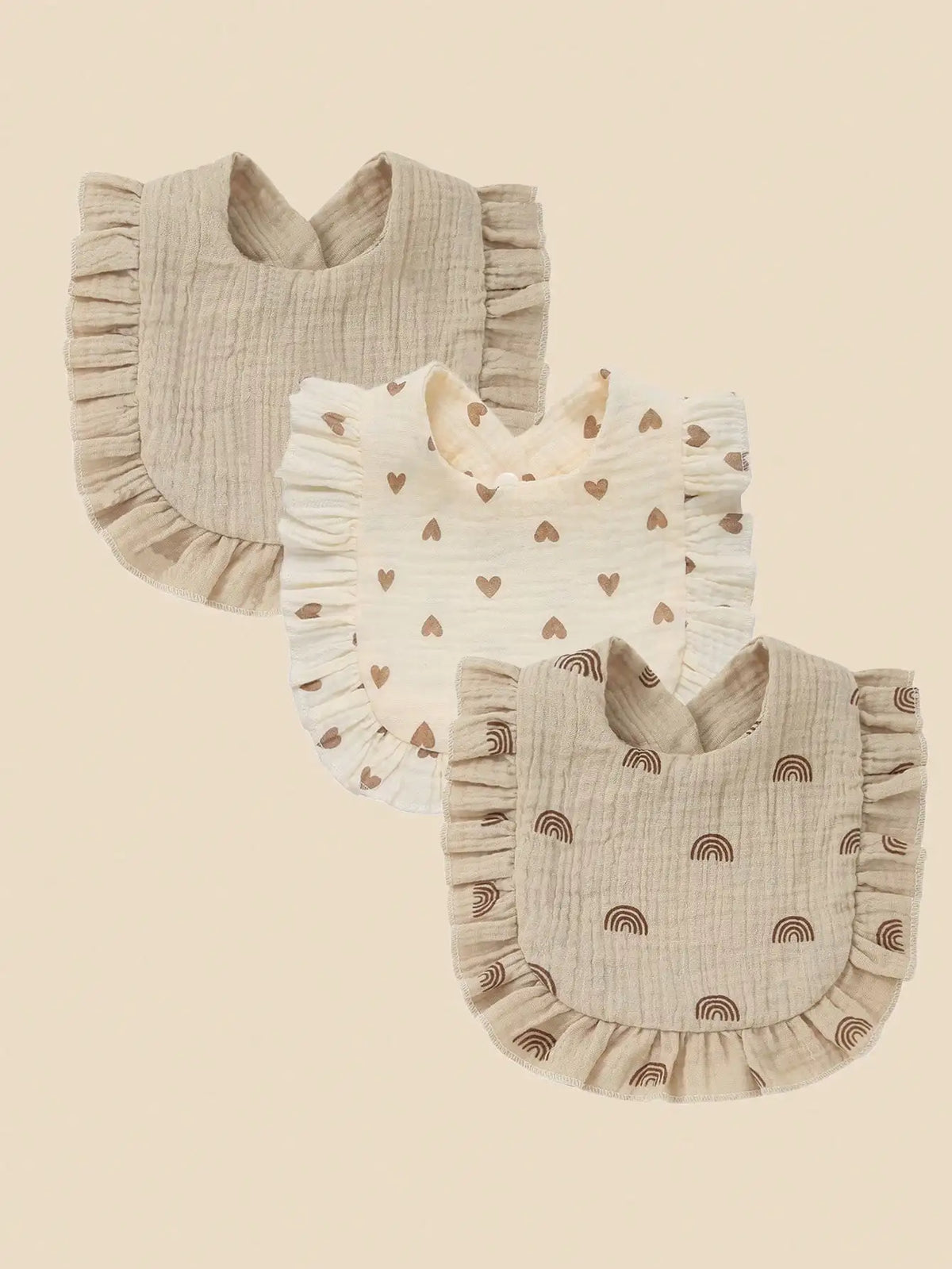 A set of three light beige cotton muslin baby bibs with ruffled edges and decorative prints.