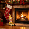 A yellow elf figure is sitting on the hearth in front of a fireplace decorated with Christmas greenery and a red stocking.