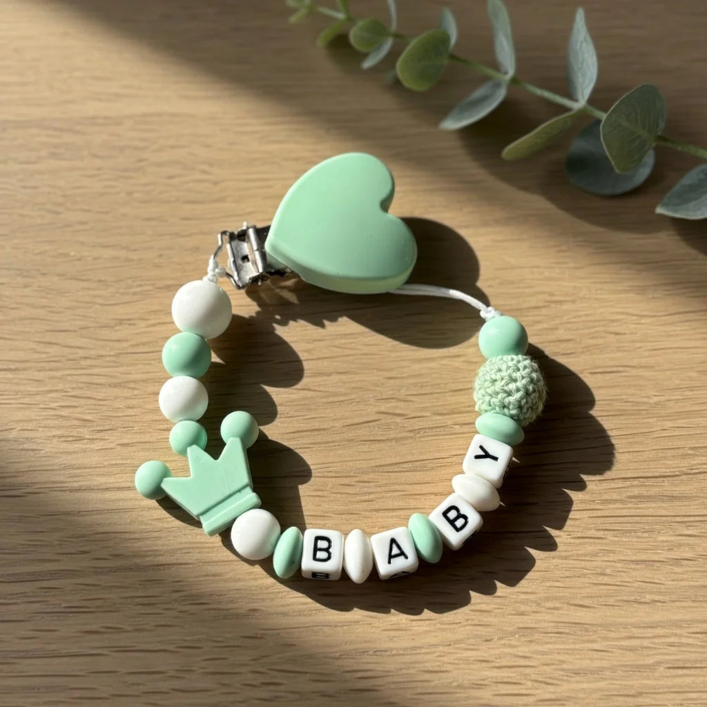 A silicone pacifier clip featuring a heart charm and a beaded bracelet with letters and a crown on a wooden surface.