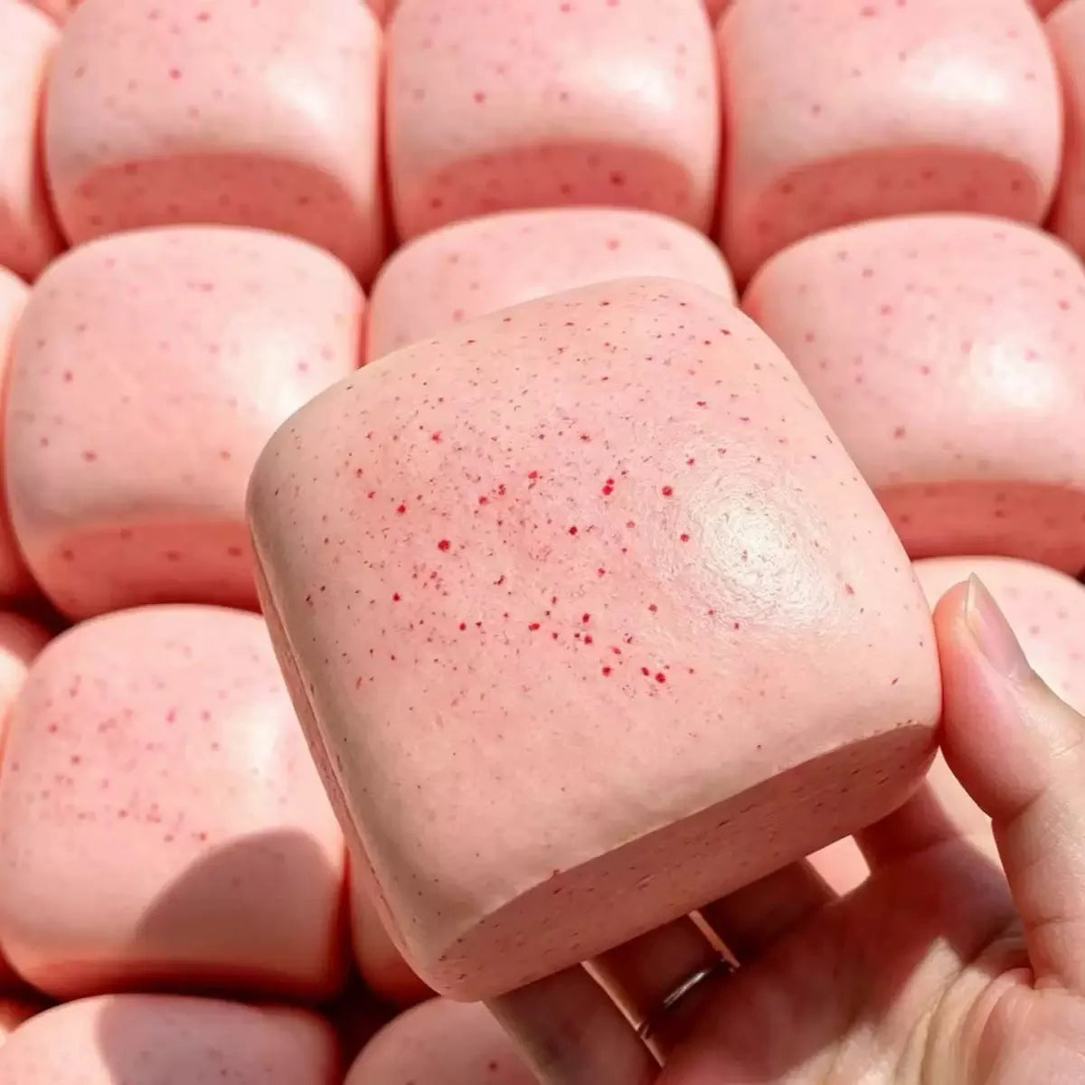 A hand holds a pink sticky cube slow-rising squishy stress relief toy with small red specks.