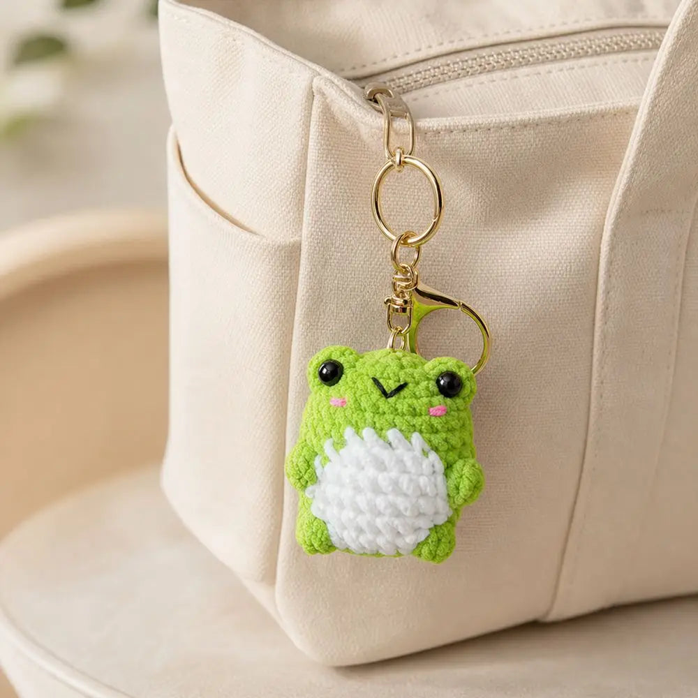 A crochet green frog charm with a white belly hanging from a gold-colored keychain attached to a light beige fabric bag.