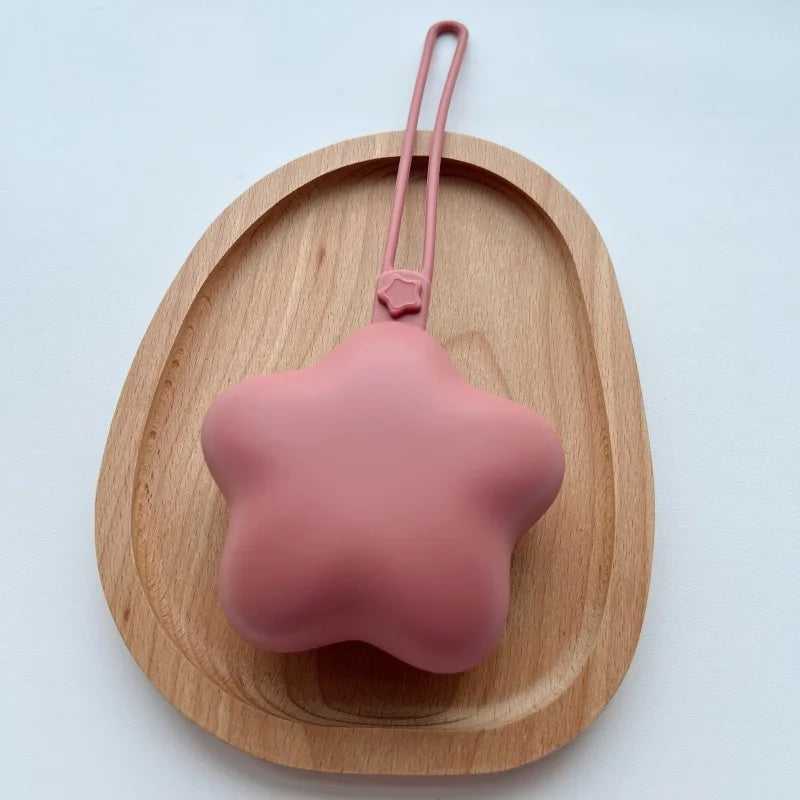 A pink, star-shaped portable pacifier and teether with a strap is displayed on a wooden tray.