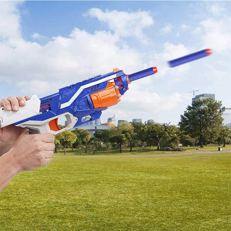 A hand holds a blue toy blaster firing a blue dart toward the sky against a backdrop of green grass and buildings.