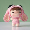 A handmade pink crochet doll with black hair and a pink bonnet against a light green background.