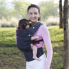 A woman wearing a pink long-sleeved shirt and white pants is holding a baby in an adjustable cotton carrier wrap outdoors.