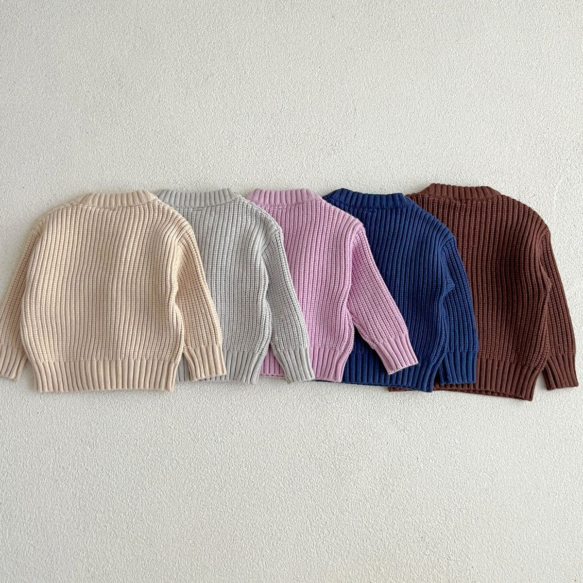 A row of nine soft cotton knit baby pullover sweaters in pastel and earthy tones laid out horizontally.