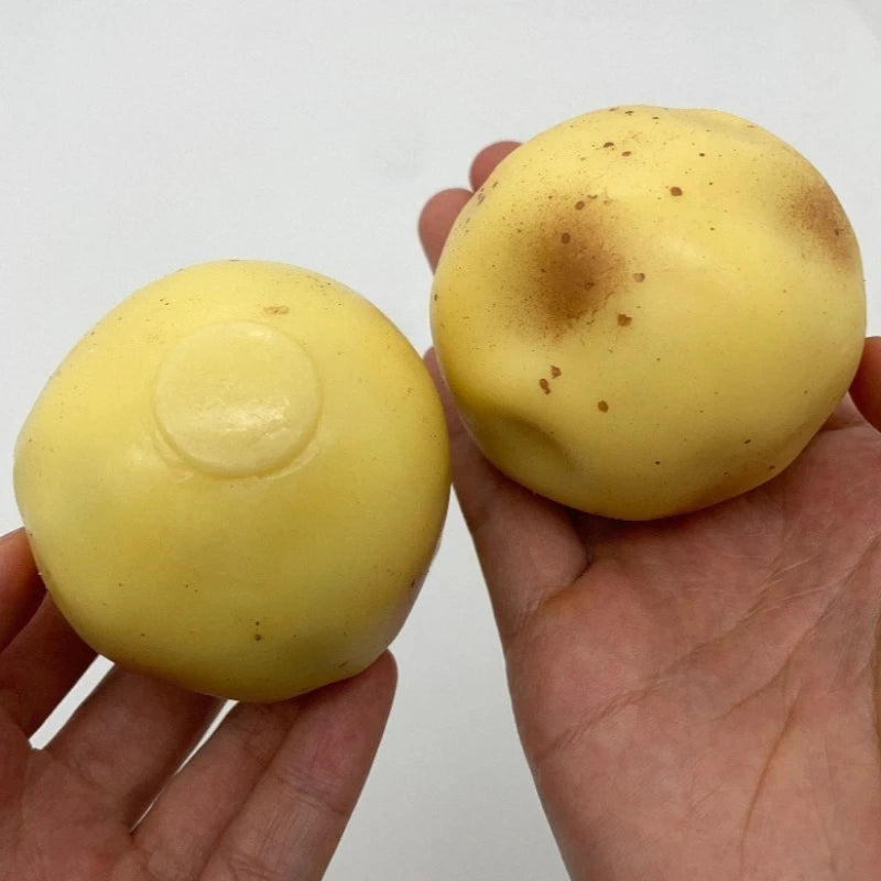 A hand holds a yellow, squishy potato stress ball with brown spots against a white background.