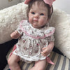 A lifelike 18-inch reborn baby doll with hand-painted details wearing a floral dress and pink bow, sitting on a white fluffy 