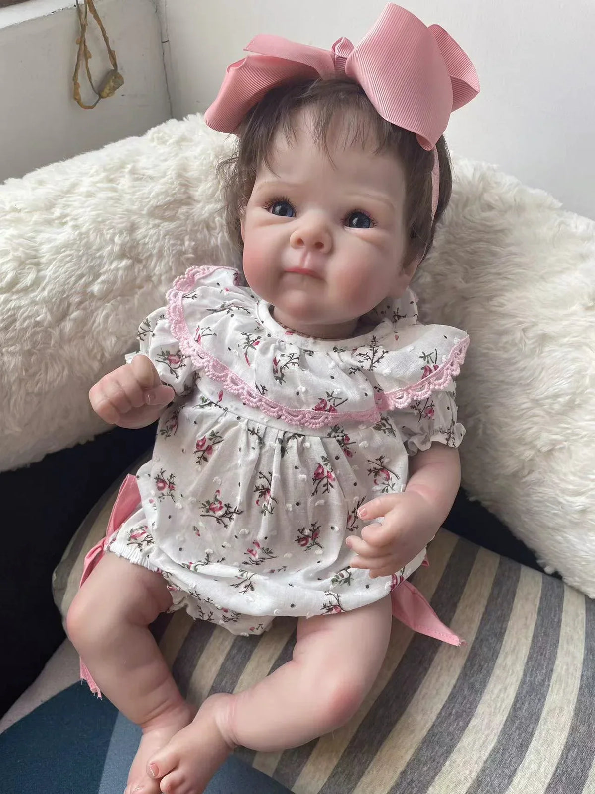 A lifelike 18-inch reborn baby doll with hand-painted details wearing a floral dress and pink bow, sitting on a white fluffy 