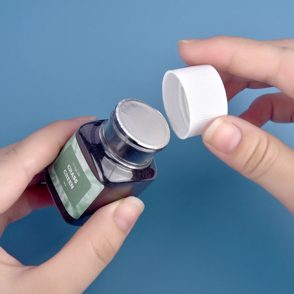 A hand holds a glass jar of green textile fabric dye powder and a white plastic cap against a blue background.