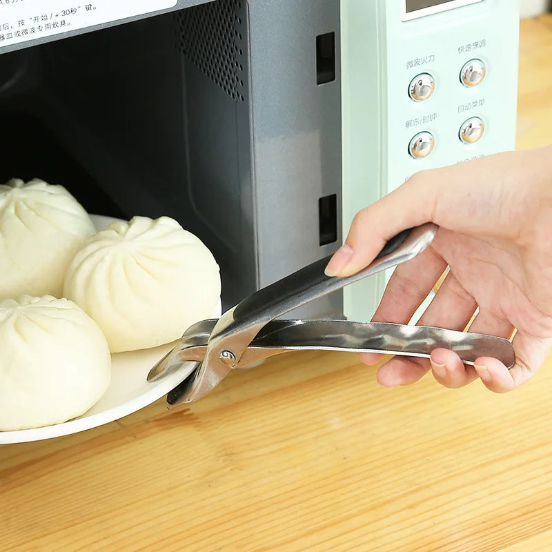 A hand uses metal tongs to grip a white steamed dumpling on a plate next to an electronic device.