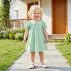 A toddler girl wearing a green plaid A-line dress with a Peter Pan collar standing outdoors.