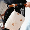 A quilted cream-colored baby diaper caddy organizer with brown leather straps is shown hanging on a stroller.