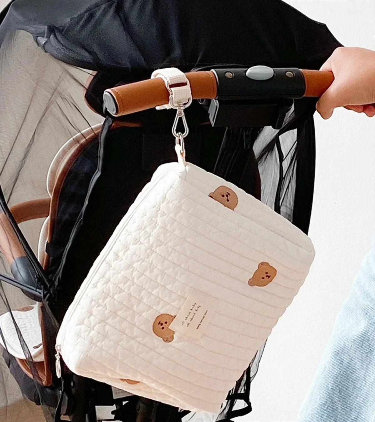 A quilted cream-colored baby diaper caddy organizer with brown leather straps is shown hanging on a stroller.
