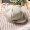 A beige knit romper and newsboy cap set displayed on a woven mat.
