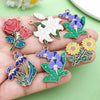 A hand holds various enamel flower charms in shades of red, white, blue, and yellow on a green background.