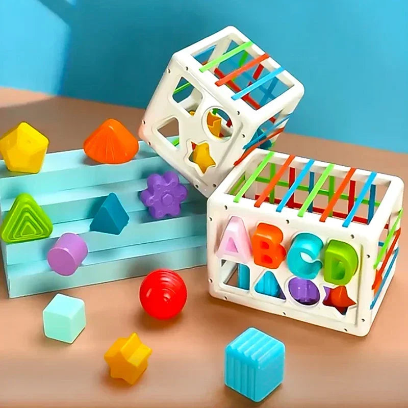 A white cube sorting toy with colored elastic bands and various geometric shapes on a wooden surface.