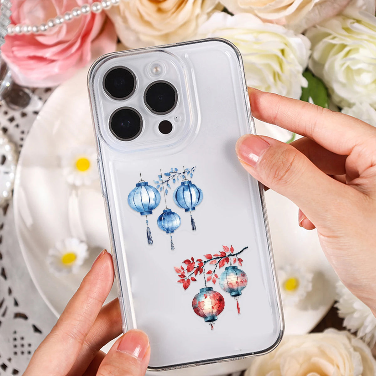 A hand holds a clear phone case featuring blue and pink lantern stickers against a background of white flowers.