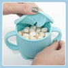 A hand is holding a light blue silicone snack cup with a lid, filled with small white balls for babies and toddlers.